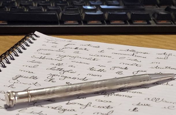Appunti scritti a mano in corsivo su un quaderno, con una penna e una tastiera del computer sullo sfondo.