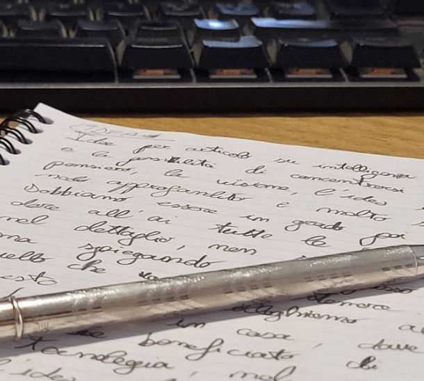 Appunti scritti a mano in corsivo su un quaderno, con una penna e una tastiera del computer sullo sfondo.