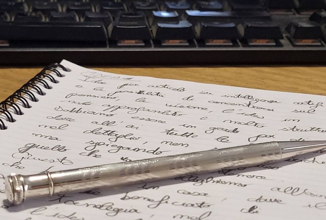Appunti scritti a mano in corsivo su un quaderno, con una penna e una tastiera del computer sullo sfondo.
