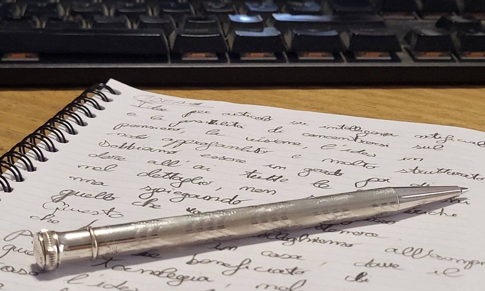 Appunti scritti a mano in corsivo su un quaderno, con una penna e una tastiera del computer sullo sfondo.
